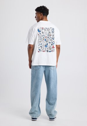 Una camiseta blanca de manga corta presenta un gráfico colorido de varios garabatos en la parte posterior, combinada con unos vaqueros de pierna ancha en un tono azul claro.