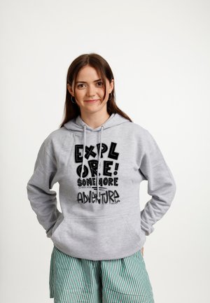 Ung kvinde iført en grå hoodie med teksten "Explore! Some More Adventure" og grønstribede shorts, står med hænderne i lommerne og smiler.
