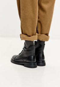 Botas de cuero negro con cordones, usadas con calcetines grises y pantalones de pana marrón arremangados, sobre un fondo blanco.