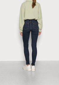 Mujer vistiendo unos pantalones skinny azul oscuro, una blusa de manga larga verde claro y zapatillas blancas, de pie contra un fondo liso.