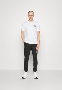 Camiseta blanca de manga corta con un pequeño logo negro en el pecho, combinada con pantalones de chándal negros que presentan texto impreso en blanco y zapatillas blancas.