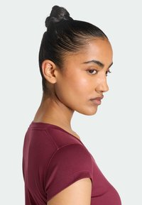 Vrouw met donker haar in een knot die een korte mouwen bordeauxrode blouse draagt, kijkt opzij tegen een effen lichte achtergrond.