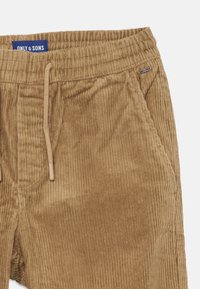 Pantalons en velours côtelé marron avec texture côtelée visible, taille élastique, fermeture à cordon de serrage et petite étiquette de marque.
