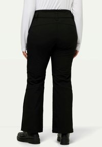 Zwarte flarebroeken met een gestructureerde tailleband, zijsplitten bij de manchetten en een gladde stof; gedragen met zwarte laarzen en een witte longsleeve top.