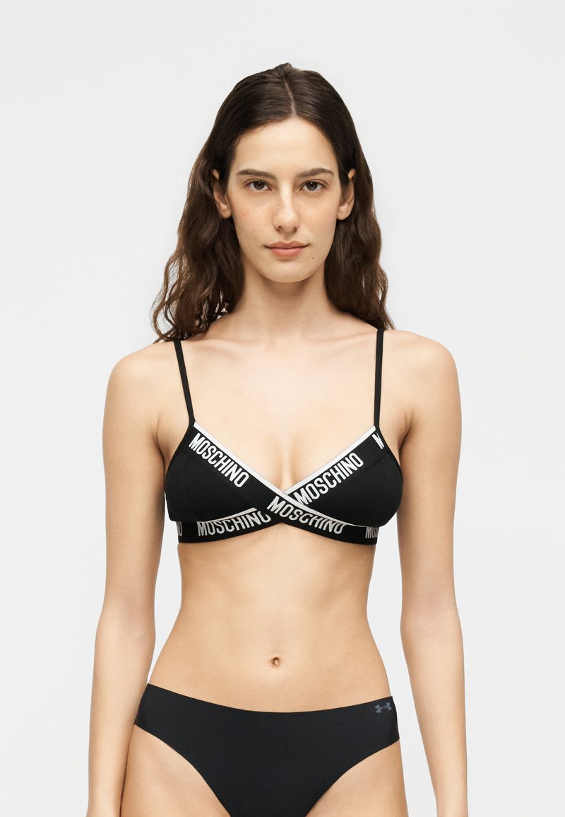 Νέα γυναίκα με μακριά καστανά μαλλιά που φοράει μαύρο τριγωνικό bralette Moschino και μαύρο κάτω μέρος μπικίνι Under Armour, στέκεται μπροστά σε λευκό φόντο.