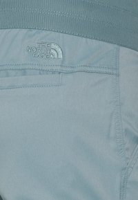 Ljusblått tyg med sydd ficka och broderad "The North Face"-logotyp nära resåren i midjan.