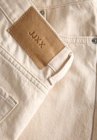 Μπεζ ύφασμα τζιν με καφέ δερμάτινο πάτο με την ετικέτα "JJXX", ραμμένο κοντά σε πίσω τσέπη και λεπτομέρεια λουριού ζώνης.
