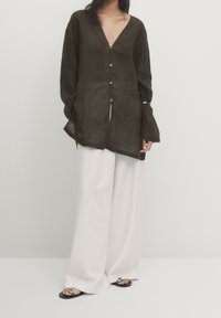 Chemise en lin vert olive foncé avec un décolleté en V profond, des boutons sur le devant et des manches longues légèrement évasées, assortie à un pantalon blanc à jambes larges et des sandales noires.