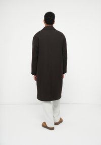 Uomo in piedi di spalle, indossa un lungo cappotto marrone scuro, pantaloni bianchi e scarpe marroni, su uno sfondo bianco uniforme.