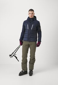 Námořnická puffer bunda s dvěma bočními kapsami, bílými zipy, nastavitelnou kapucí, olivově zelené kalhoty, černé boty a lyžařské hole. Rukavice jsou vínové.