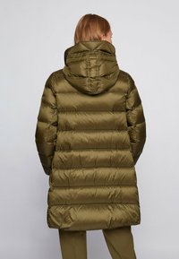 Zelený olivový prošívaný kabát s kapucí, s horizontálním prošíváním, lesklou texturou a délkou do poloviny stehen. Pohled zezadu ukazuje elegantní siluetu.
