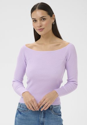 Mlada ženska v rebrastem, odkritih ramenih vijoličnem puloverju in modrih kavbojkah, stoji z rokami nežno prepletenimi spredaj.