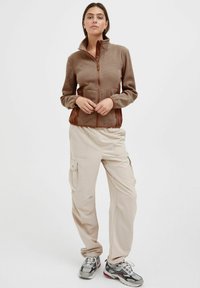 Brown zip-up fleece jacket with textured fabric and high collar, paired with beige cargo pants and grey sneakers featuring red accents.