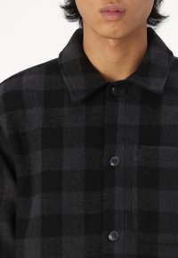 Chemise en flanelle à carreaux noirs et gris avec col, poche avant et boutons noirs, présentant une finition douce et texturée.
