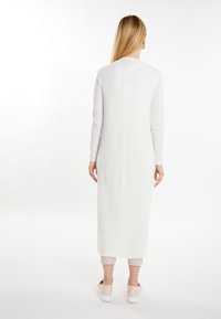 Rochie vest din tricot alb, cu un fason lung și fluid, cu fante laterale. Poartă peste un top cu mânecă lungă și încălțăminte ușoară.