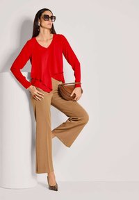 Frau trägt eine rote Bluse, beige weite Hosen, Absatzschuhe mit Leopardenmuster, große Sonnenbrille, hält eine braune Handtasche und lehnt an einem weißen zylindrischen Podest.