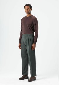 Bruine trui met lange mouwen, groene geplooide broek, donkerbruine leren schoenen, met knoop- en naaddetails, tegen een lichte achtergrond.