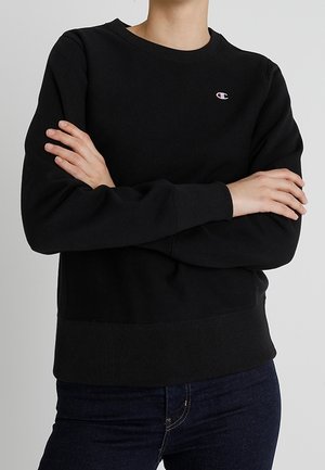 Persoon draagt een zwart Champion sweatshirt met lange mouwen, armen gekruist, gecombineerd met donkerblauwe jeans.
