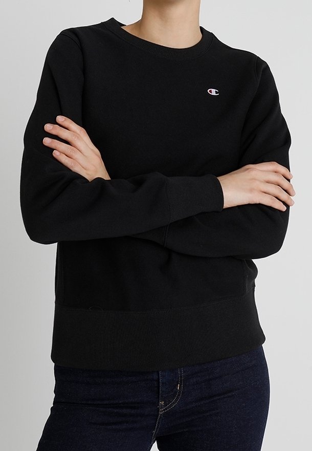Personne portant un sweat-shirt Champion noir à manches longues, les bras croisés, associé à un jean bleu foncé.