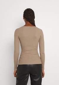 Lichtbruine geribbelde longsleeve top met een aansluitende snit en ronde halslijn, voorzien van een klein logo aan de achterkant. Gepaard met zwarte broeken.