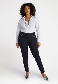 Camicia bianca a maniche lunghe con bottoni, jeans skinny scuri con orli sfrangiati, collana d'argento e tacchi alti metallici.