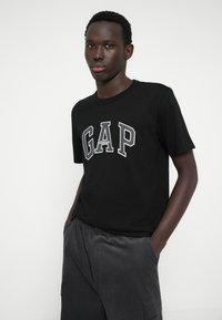 Czarna koszulka z krótkim rękawem z logiem "GAP" w odważnych białych literach na piersi, w połączeniu z szarymi spodniami sztruksowymi. Gładka tekstura materiału.