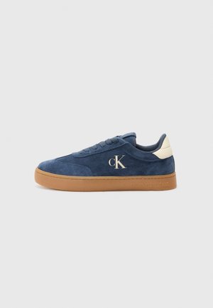 Blå ruskind lavtop sneaker med tan gummisål, hvid hælfane og "cK" logo på siden.