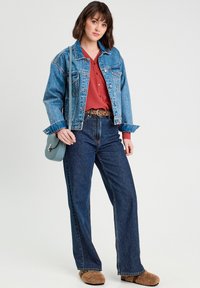 Veste en denim bleu clair, chemise rouge à boutons, jeans taille haute bleu foncé, pantoufles brunes en peluche, et un petit sac bandoulière bleu.