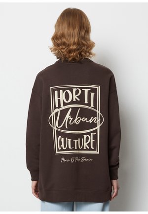 Personne aux cheveux ondulés mi-longs portant un sweat-shirt marron foncé avec "HORTI Urban CULTURE" et "Marc O’Polo Denim" imprimés dans le dos.
