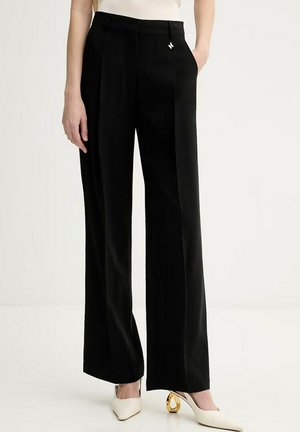 Donna che indossa pantaloni neri a gamba larga a vita alta con un piccolo ciondolo metallico sulla fascia della vita, abbinati a tacchi bianchi con tacco dorato scolpito.