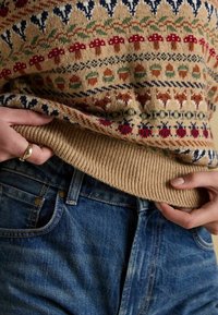 Kudumisvärk, millel on beež aluspind ja mitmevärvilised mustrid. Ribitud äär, kombineeritud siniste teksadega, millel on detailne õmblus.