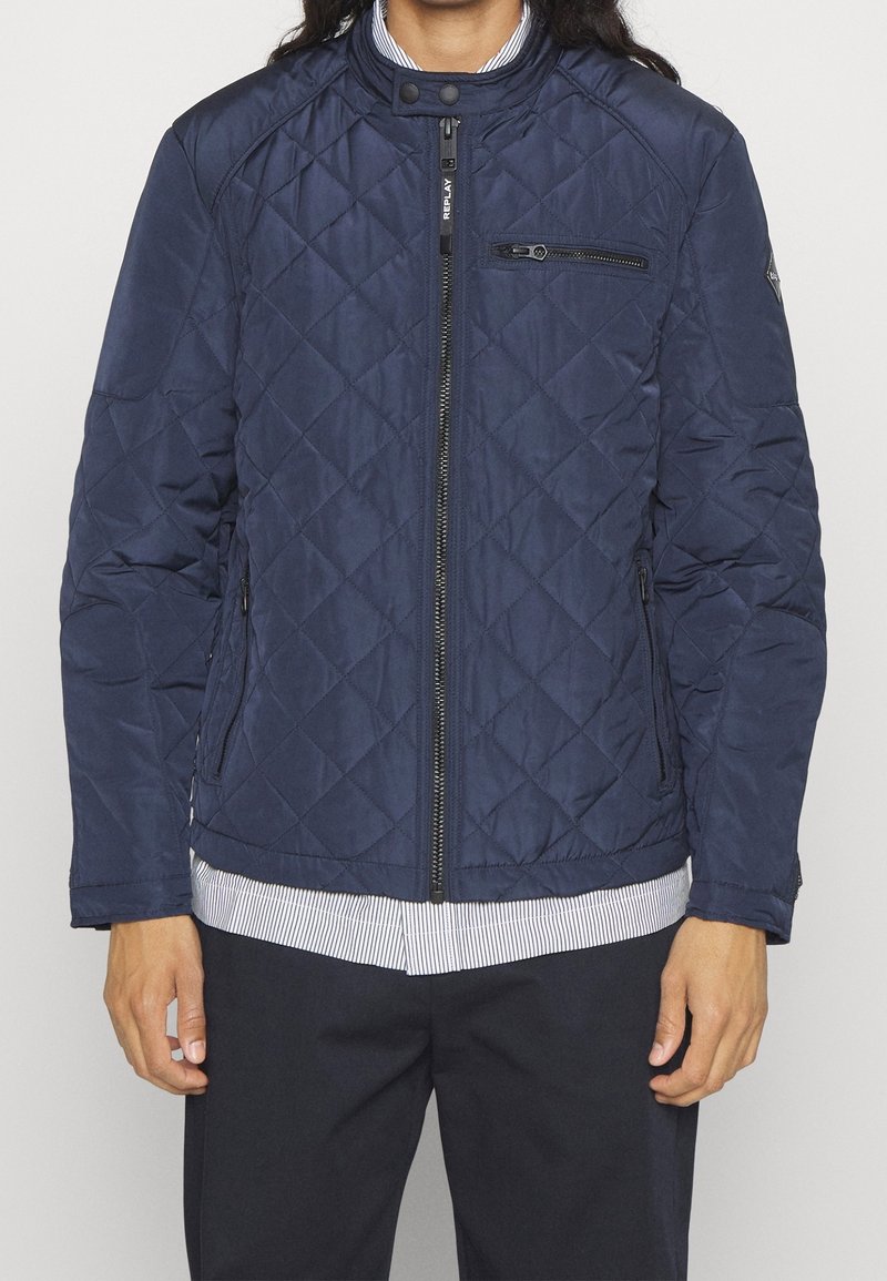 Personne portant une veste matelassée bleu marine zippée sur une chemise rayée boutonnée et un pantalon noir, debout devant un fond uni.