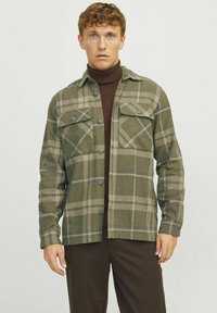 Geruite flanellen shirt in olijfgroen en beige, met twee borstzakken, knoopsluiting en lange mouwen. Draagt over een bruine coltrui.