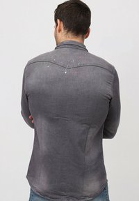Chemise en denim gris, à boutons et à manches longues, avec une finition légèrement délavée et des éclaboussures de peinture sur le haut du dos.