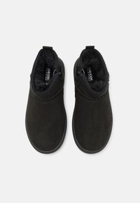 Botas de tobillo de ante negro con un suave forro de piel de pelo y cremalleras laterales. Las suelas tienen una textura antideslizante, con una forma redondeada y costuras mínimas.