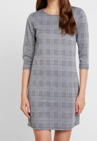 Harmaa ruudullinen mekko, jossa on pyöreä kaula-aukko ja kolmivartiset hihat, valmistettu teksturoidusta kankaasta, jossa on klassinen houndstooth-kuvio.