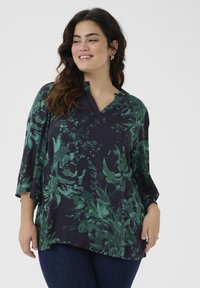 Blouse floral bleu foncé avec motif de feuillage vert, manches 3/4, décolleté en V, et texture de tissu lisse, associée à un jean foncé.