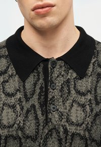 Άνδρας που φοράει πλεκτό πόλο πουκάμισο με κουμπιά, μαύρο γιακά και αφηρημένο μαύρο μοτίβο σε γκρι ύφασμα.