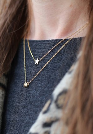 Goldene Halsketten übereinander geschichtet; eine hat einen Stern-Anhänger und die andere einen Blockbuchstaben "M". Getragen über einem dunklen, strukturierten Pullover.