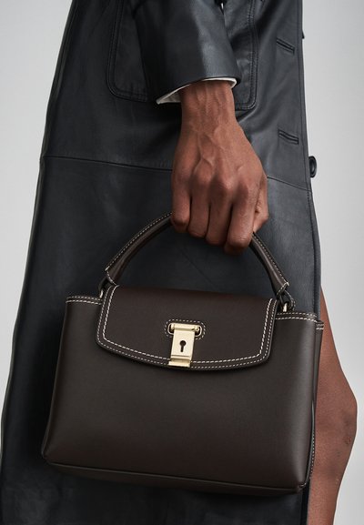 Brown leather handbag with a structured shape, gold hardware lock, and white stitching accents. The handle is short and rounded.