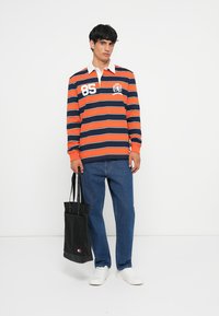 Polo de manga longa riscado em laranja e azul marinho com gola branca, apresentando um gráfico de leão e o número 85. Combinado com jeans azuis e uma bolsa tote preta.