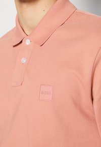 Polo rose en tissu texturé avec un col côtelé, présentant un petit patch logo carré en bas à gauche.