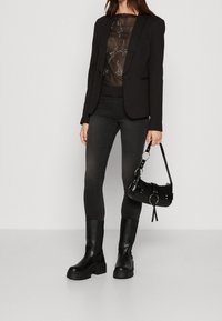 Svart kavaj över en genomskinlig topp, kombinerad med mörka slim fit-jeans och kraftiga svarta ankelstövlar. Accessoarer inkluderar en liten svart väska med detaljer.