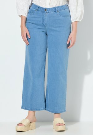 Kobieta w jasnoniebieskich szerokich dżinsach z elastycznym pasem i plecionych sandałach na platformie, stojąca na białym tle.