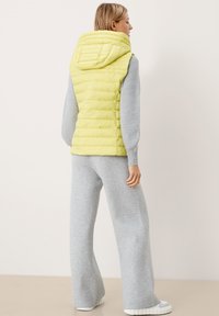 Gelbe gepolsterte Weste mit Kapuze, ausgestattet mit horizontalen Steppnähten. Kombiniert mit grauen, weit geschnittenen Sweatpants. Weicher Stoff und lässiges Design.