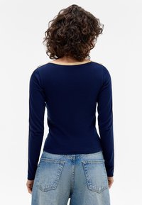 Marineblå langermet topp med rund hals og kontrasterende beige skulderdetaljer, laget av mykt, elastisk stoff. Brukt med lyseblå jeans.