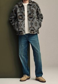 Fleecejacke mit grau-braunem Muster, kombiniert mit blauer Jeans und hellbraunen Schuhen, vor einem neutralen Hintergrund stehend.