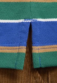 Gros plan sur l’ourlet fendu d’un vêtement en tricot à rayures horizontales vertes, bleues, beiges et blanches sur du tissu en velours côtelé marron.