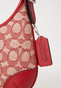 Borsa a mano rossa con tessuto intrecciato e motivo a cerchi ripetuti, dettagli in pelle e un'etichetta metallica con scritto "Coach."