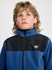 Blond dziecko z piegami, ubrany w niebiesko-czarną polarową kurtkę na zamek z logo pszczoły, patrzy lekko w górę na jednolitym tle.
